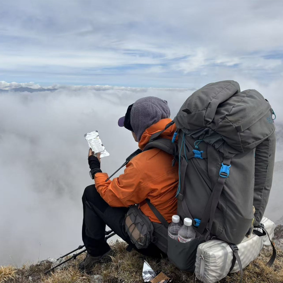 A hiker sitting on a mountain ridge above a sea of clouds, wearing an orange jacket, black pants, and a large gray backpack while holding a packaged PEAK4RATION Emergency Food Ration Bar.