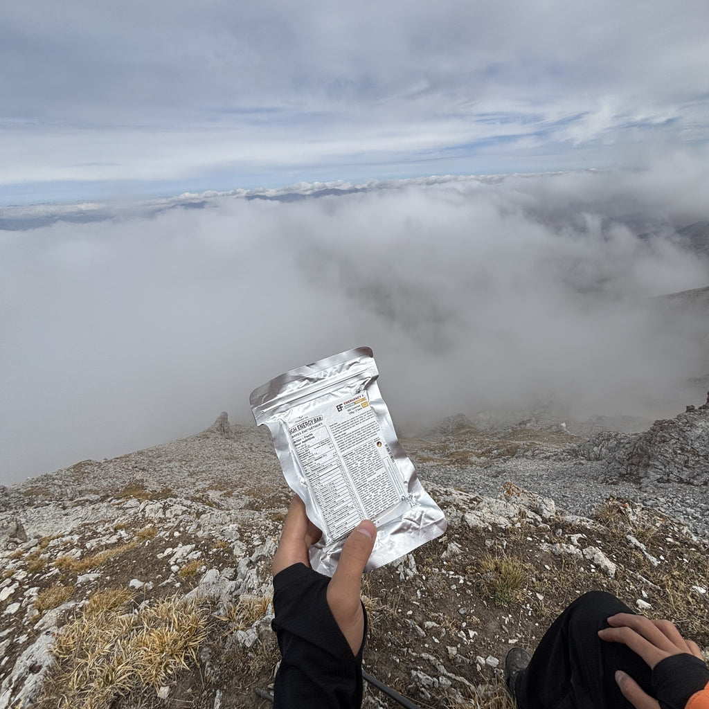 A person on a rocky mountain peak holds up a PEAK4RATION Emergency Food Ration MRE Bar, showing the back of the package with printed text, with a sea of clouds and mountain landscape in the background.