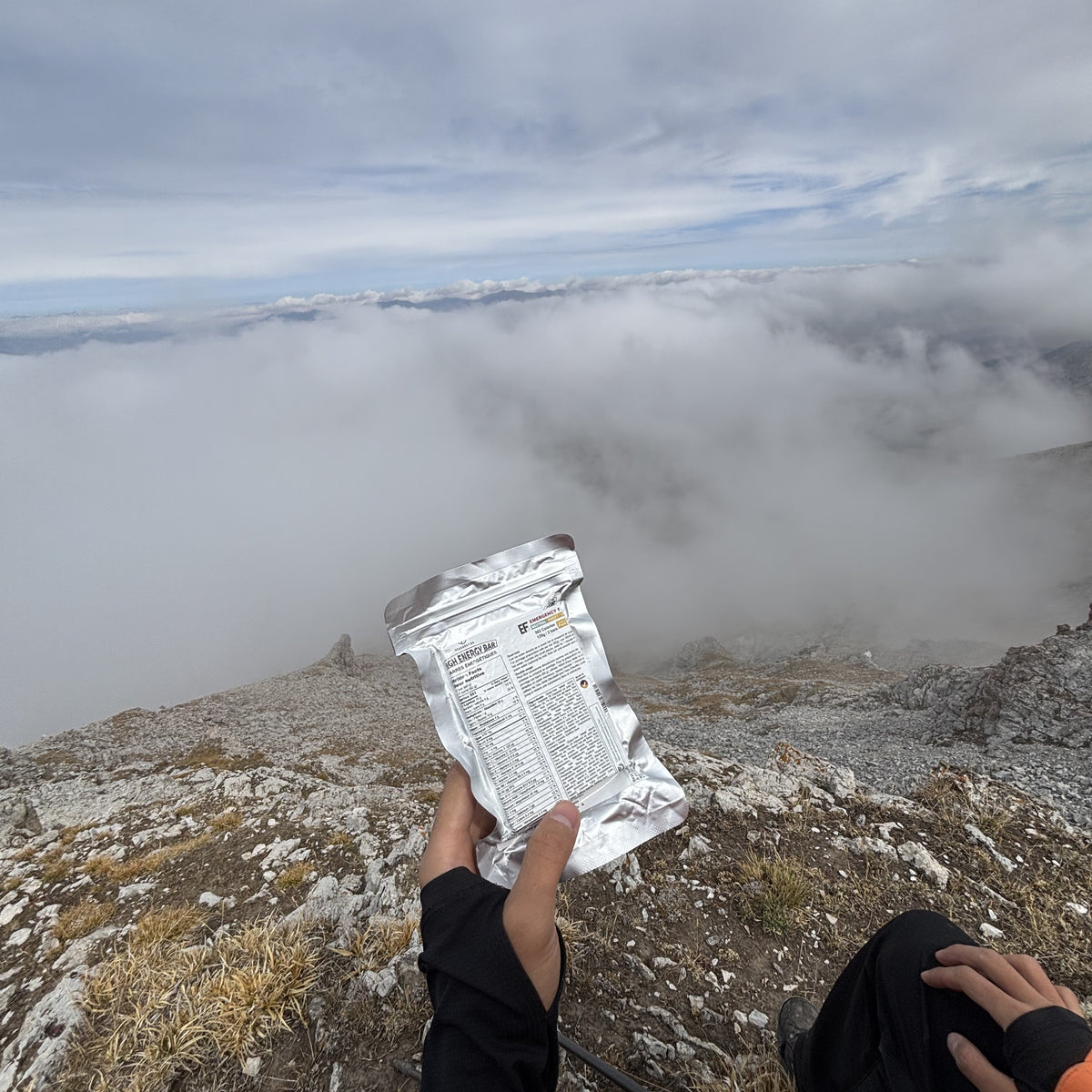 A person on a rocky mountain peak holds up a PEAK4RATION Emergency Food Ration MRE Bar, showing the back of the package with printed text, with a sea of clouds and mountain landscape in the background.