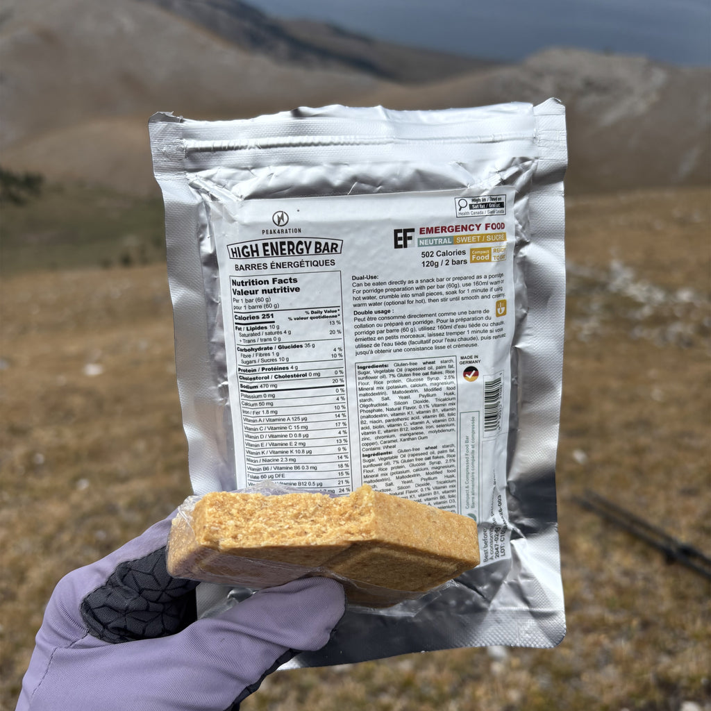 A hand wearing a purple glove holds a silver pouch of PEAK4RATION Energy Bar emergency food and a square piece of the golden-brown baked bar in front of a blurred outdoor mountain landscape.